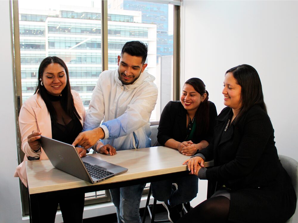 Cuatro colegas discutiendo y sonriendo alrededor de una computadora portátil en una oficina.