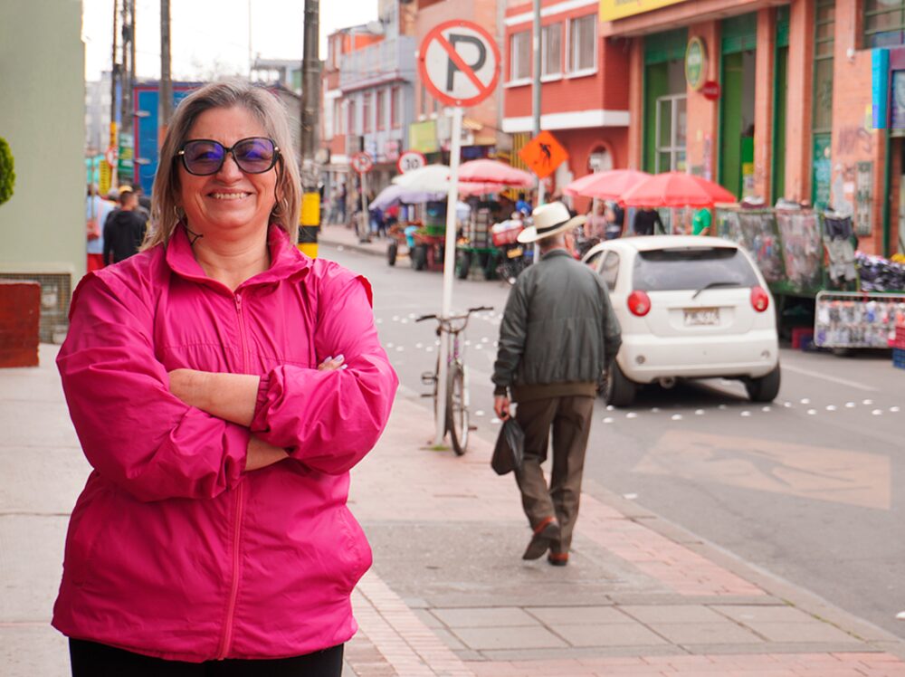 Mujer con chaqueta rosa sonriendo en una calle muy transitada.