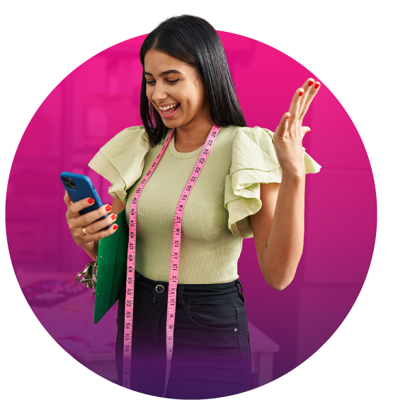 Una mujer sonriente con cabello largo y oscuro sostiene un teléfono y levanta la mano. Tiene una cinta métrica rosa alrededor del cuello y está vestida con un top beige y pantalones negros.