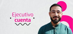 Un hombre con gafas y barba se encuentra de pie sonriendo sobre un fondo blanco y rosa con el texto "Ejecutivo cuenta".